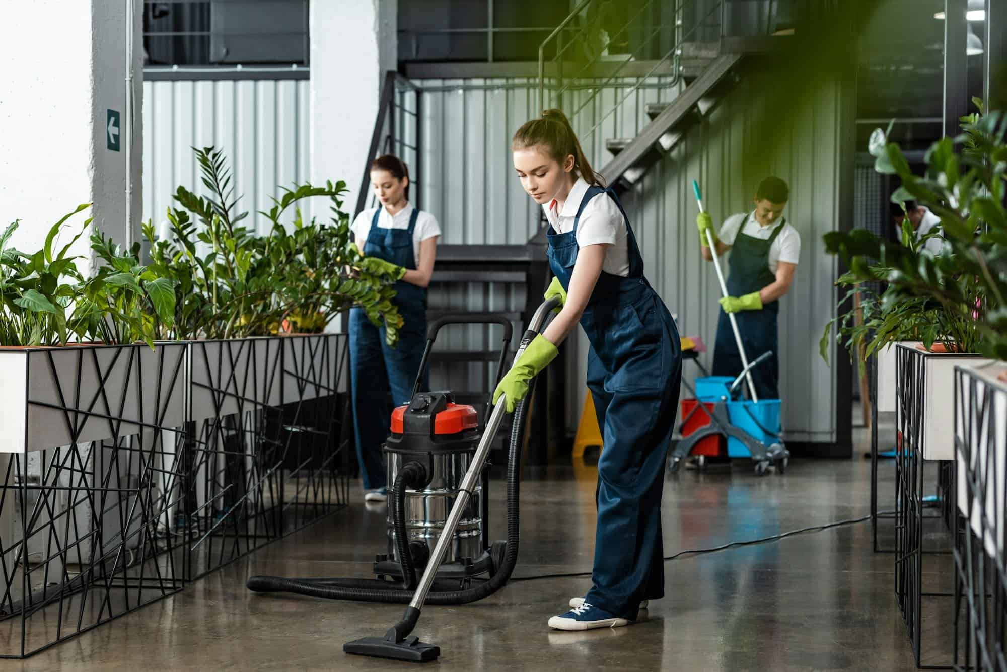 young cleaner vacuuming floor in office near colleagues